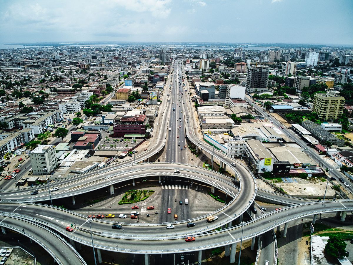 The Boulevard Valery Giscard d'Estaing interchange in Abidjan from the air, traffic looping through a raised roundabout with the Plateau towers and the Ebrie lagoon behind it