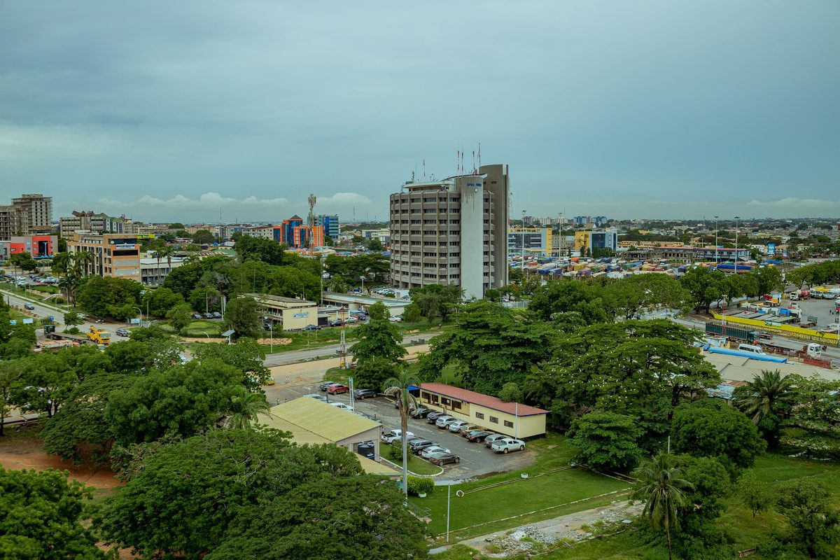 Accra from above on an overcast afternoon, a tall office block among low-rise streets, heavy tree cover through the middle of the city and stacked shipping containers on the right