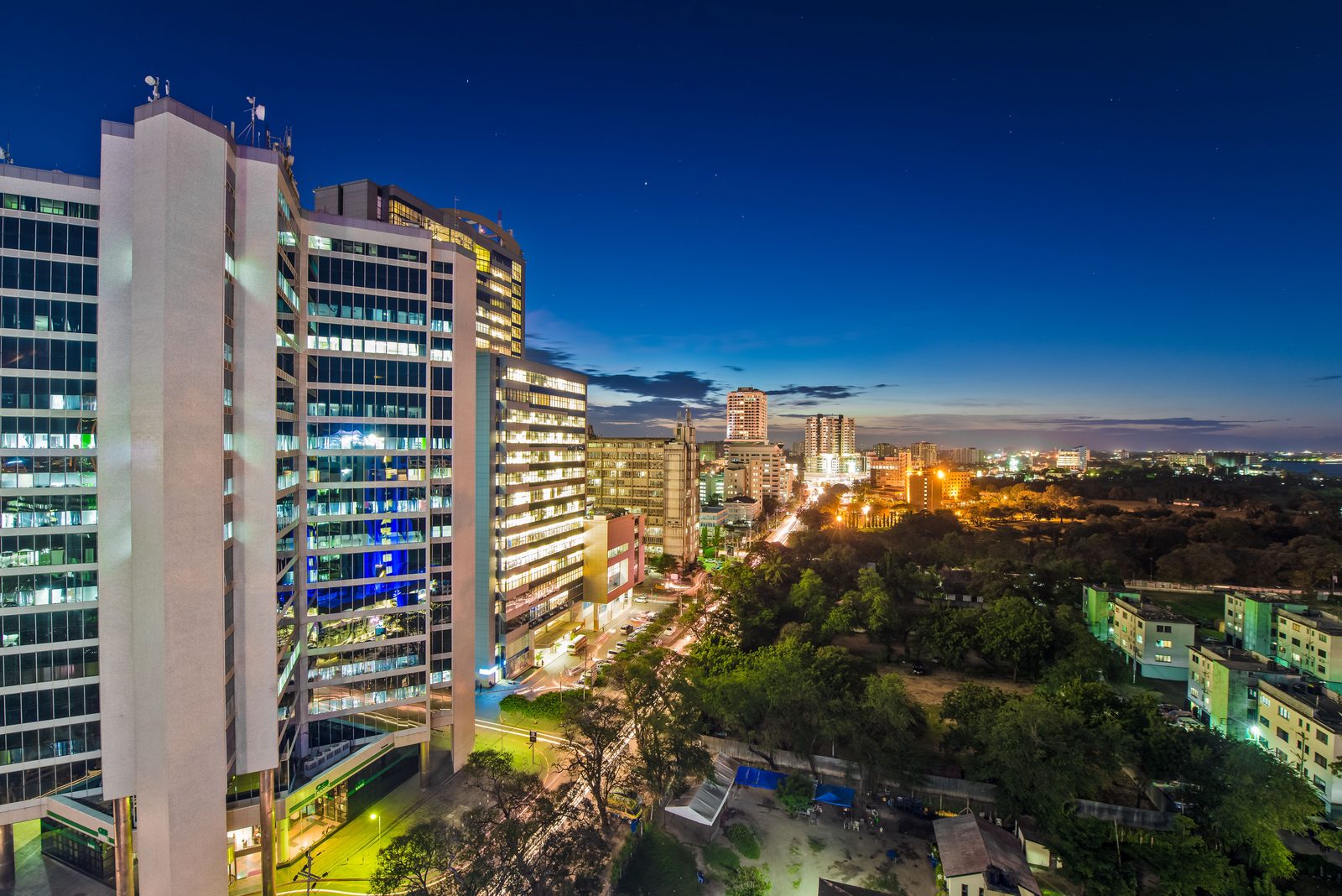 Dar es Salaam at blue hour from above: a tall pale modernist block in the foreground with its windows lit, a lit avenue running past a dark wooded park, and the harbour water and the lights of the far shore beyond it