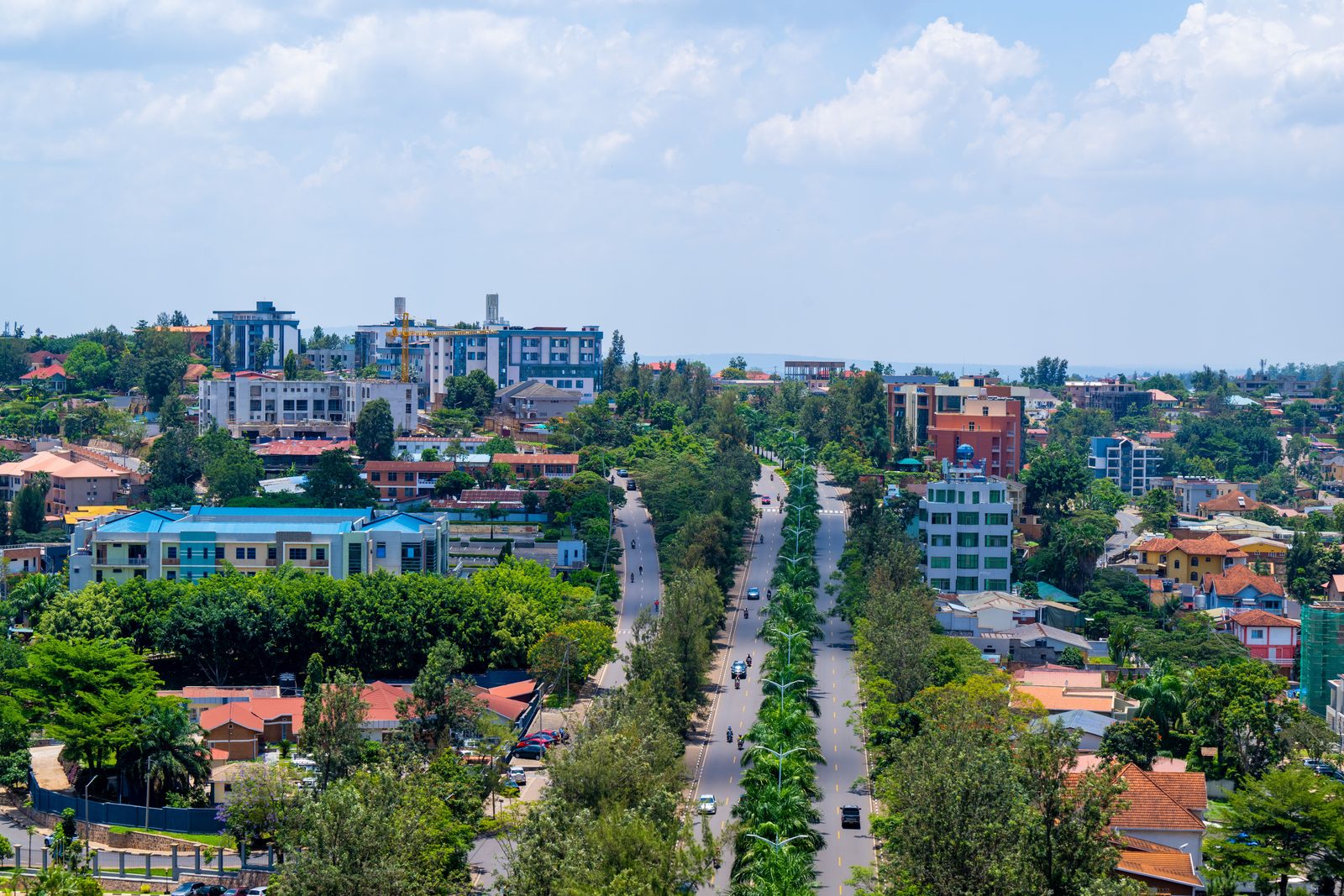 Kigali from above: a tree-lined divided boulevard running between green ridges of red-roofed houses, motorcycles and light traffic on it, the hills of the city rising on both sides
