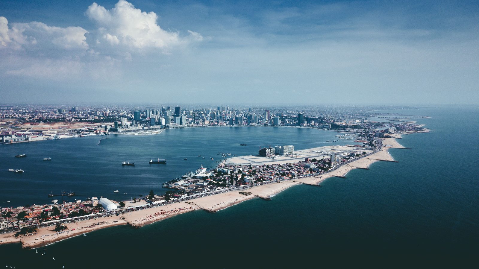 Luanda from the air: the long sand spit of the Ilha curving across the bay, ships at anchor inside it, and the towers of the city standing along the far shore