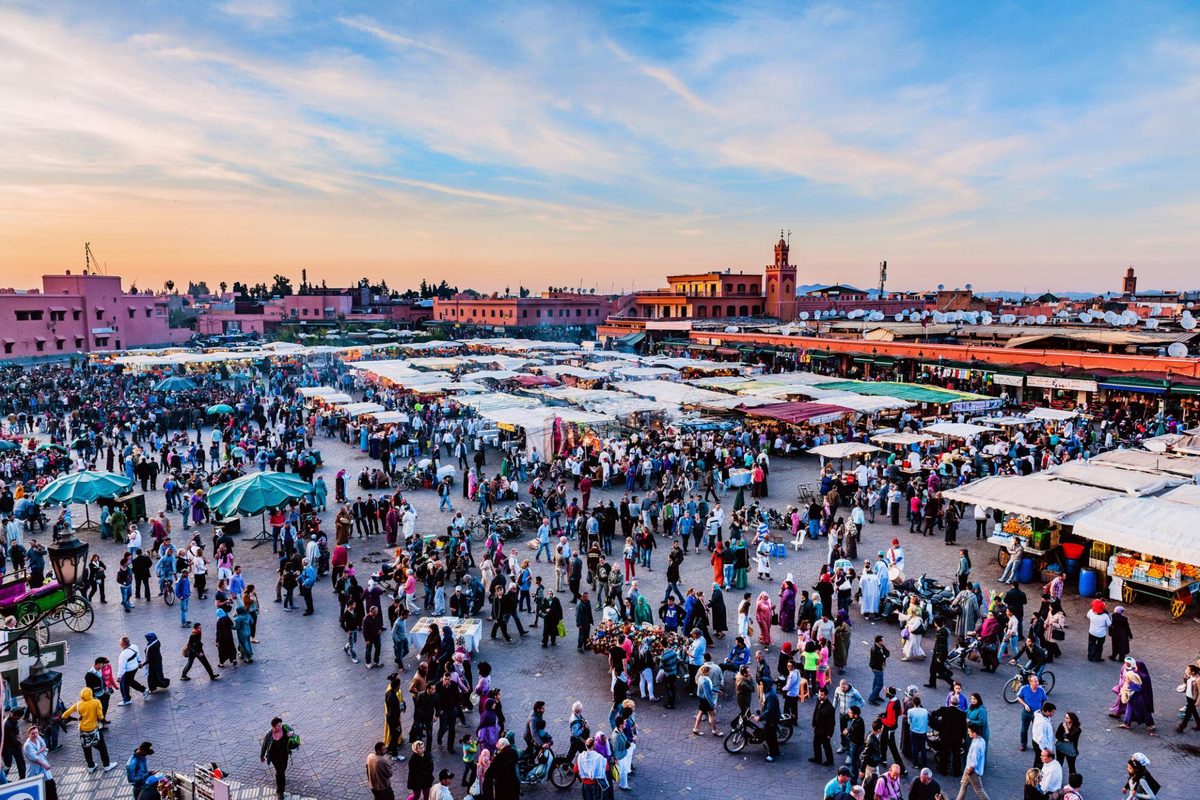 Jemaa el-Fna square in Marrakech at dusk, crowds moving between rows of covered food stalls, the pink walls of the medina and a minaret behind, with the sky going orange
