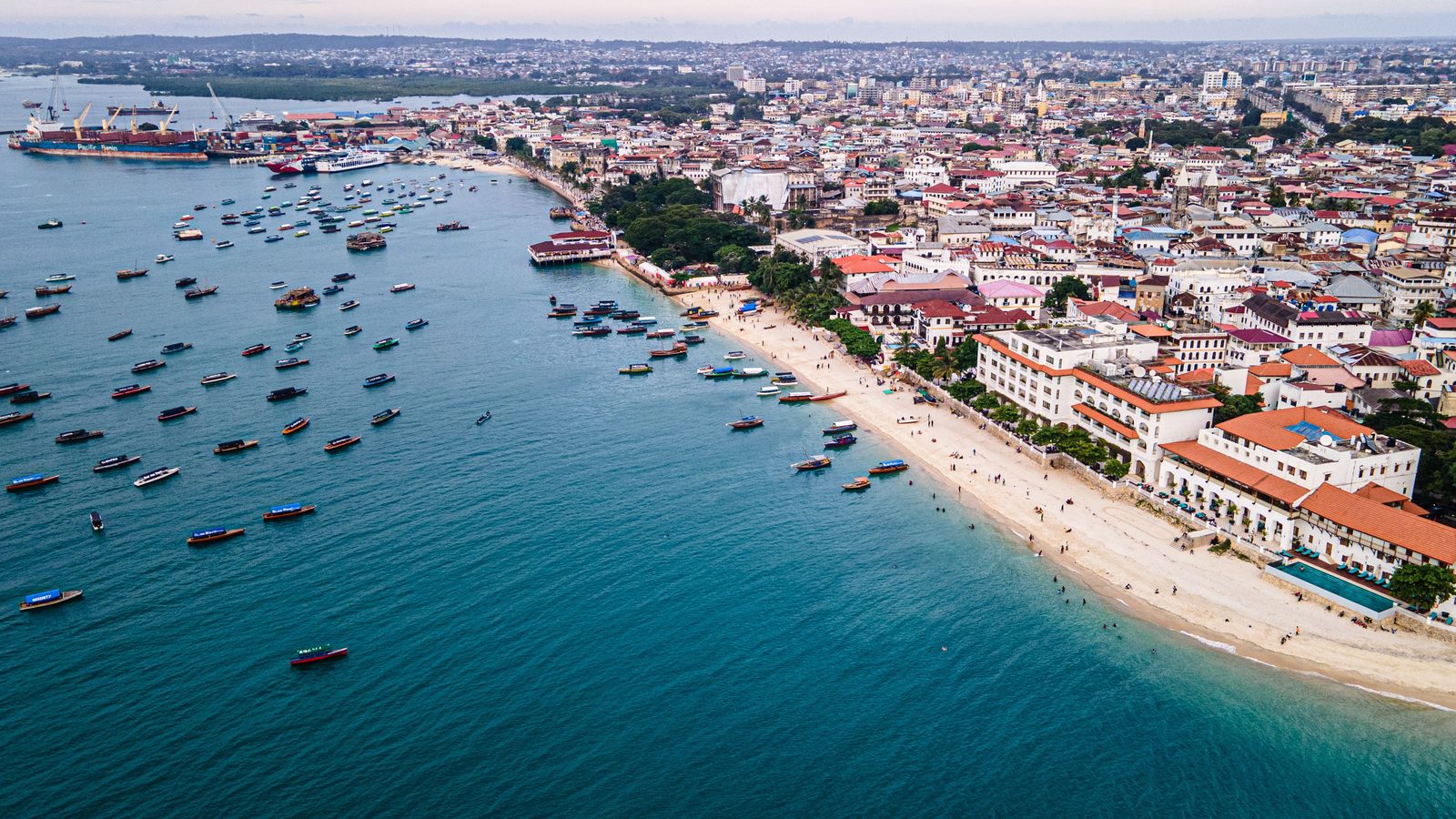Zanzibar City from the air along its waterfront: dhows and small boats moored on pale shallow water, and the low white and red-roofed buildings of Stone Town running behind the shore