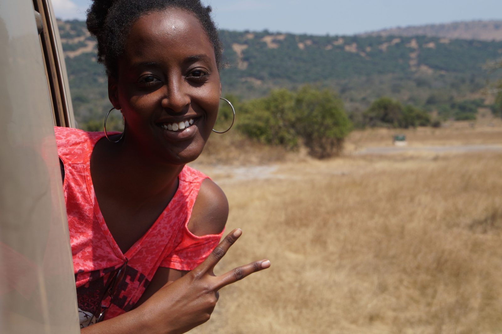 A woman leaning out of a moving vehicle's window smiling at the camera, dry hills and grassland running away behind her