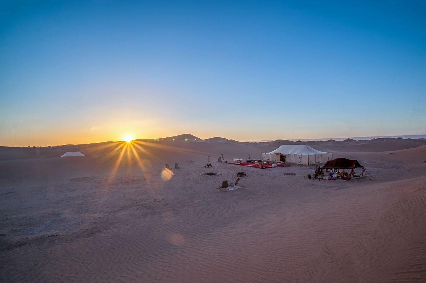 A camp of white canvas tents and a low striped awning pitched on open sand, with the sun just clear of the dunes on the horizon behind them
