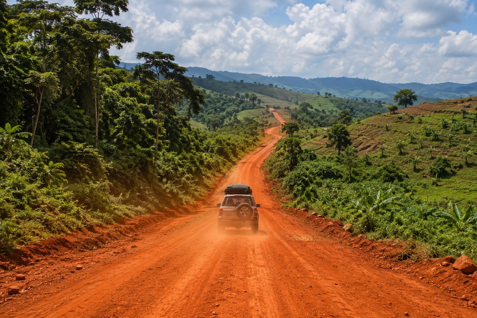 A loaded four-wheel drive with a roof box driving away on a red laterite road, dust rising behind it, the road running on over green hills