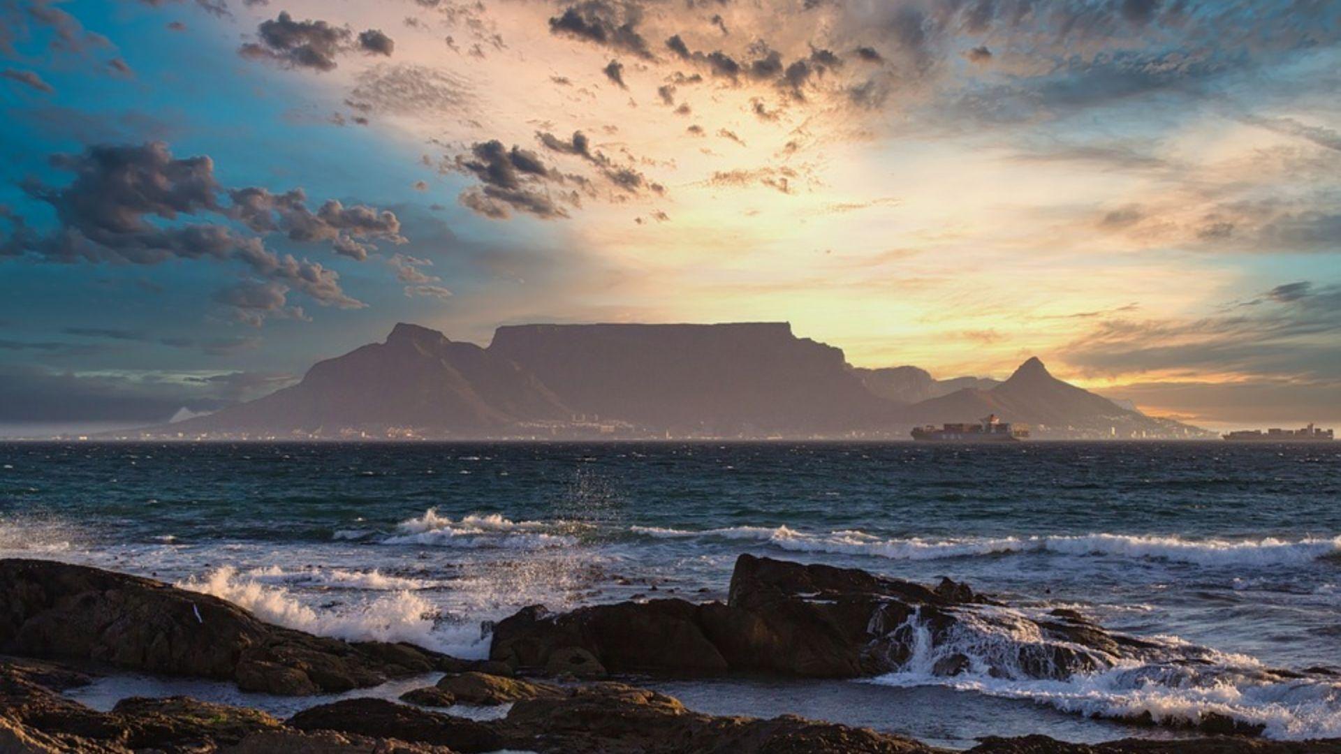 A flat-topped mountain and its two flanking peaks seen across a bay at sunset from rocks in the foreground, with surf breaking and a container ship at anchor